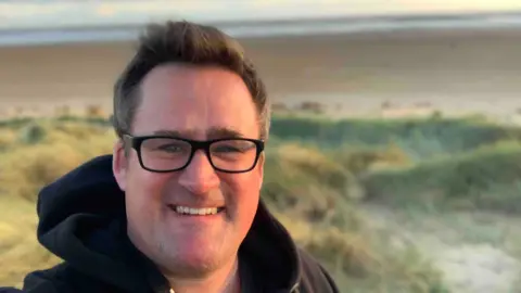 A selfie of Tom Morris wearing a black hoodie and black rectangular glasses. He is standing on sand dunes at sunset, smiling at the camera. In the distance you can see the beach and the sea.