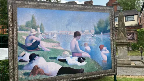 A print of a very large painting stands on a frame. The painting depicts people lay and sat by a river, with some other people in the water.