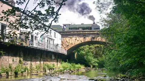 A green steam train crosses a stone arch bridge above a shallow rocky stream, with houses and dense greenery surrounding the scene.