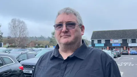 A man with dark tinted glasses and a blue shirt standing in a car park. He has grey hair and stubble. There are cars in the background and some small businesses.