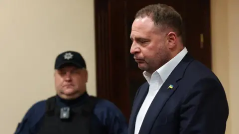 A man in a suit stands in court while a policeman looks on