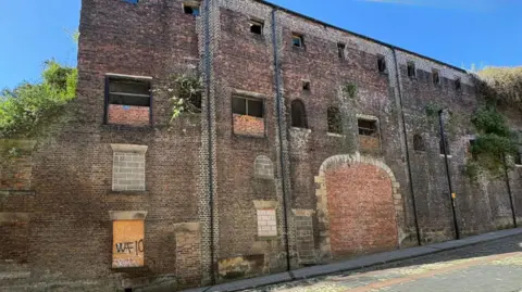 The Coppersmith's Shop. The abandoned building has trees and plants growing out of it. Many of the windows have been bricked or boarded up.