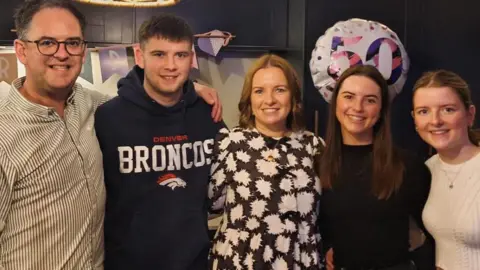 Nicola King Lucy pictured linking arms with her family. 
Nicola King is wearing a white patterned shirt and Lucy is wearing a black top. Thomas King, Lucy's brother, is wearing a Denver Bronco's hoodie and their father, Richard is in a striped shirt. Lucy's sister, Olivia, is wearing a white top and necklace.  There is a balloon with the number 50 floating behind them.