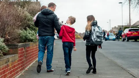 Father with 3 kids walking along the pavement, walking away from the camera. They are on a residential street in the UK 