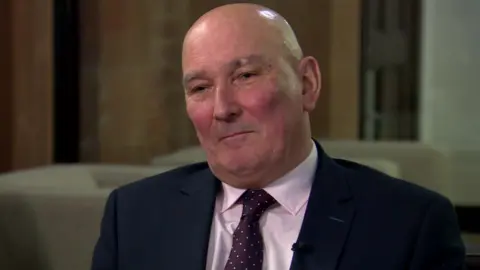 Garry Matthewson is sitting on a chair. He's wearing suit with a white shirt and a maroon spotty tie. 