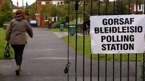 A woman arrives at a Bowls Pavillion set up as a polling station in Cardiff. She is wearing a brown coat, dark trousers and brown boots. She is walking next to a metal gate with a sign on it which reads 'polling station'.