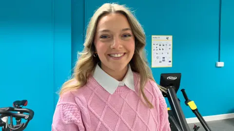 Holly is wearing a pink jumper over a white shirt. She is smiling at the camera. She has blond long hair. 