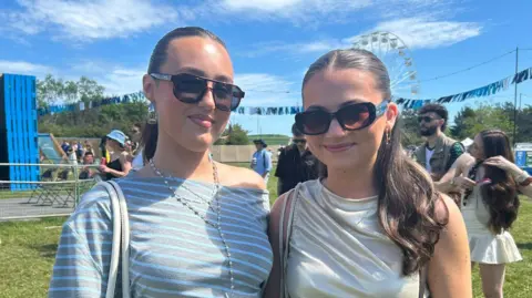 Two young women in sunglasses smile at the camera on a music festival site.