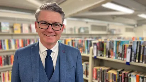 Ann Gannon / BBC Carl Ward is seen wearing a blue suit, waistcoat and tie standing in a school library, with rows of bookshelves filled with fiction and non-fiction titles behind him.