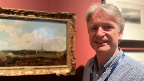 Adrian Green smiles at the camera - he wears a blue check shirt with Salisbury Museum lanyard. Blurred beside him is the painting in an ornate gold frame.