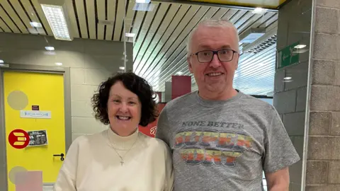 Easterhouse Phoenix Tony Deeney stands next to his wife, Teresa. Tony is wearing a grey t-shirt and black square glasses to the right of the image. He is smiling while looking at the camera. Teresa has short brown hair wears a cream jumper and stands to the left of the image. She is smiling at the camera.