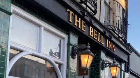 The front of The Bell Inn in Cheltenham, a pub with a green brick exterior and a black sign with gold lettering