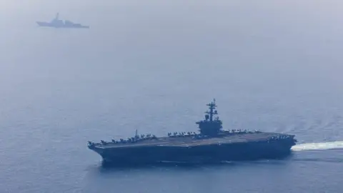 Reuters A large aircraft carrier, carrying many fighter jets, in a clear blue sea with another ship in the distance.