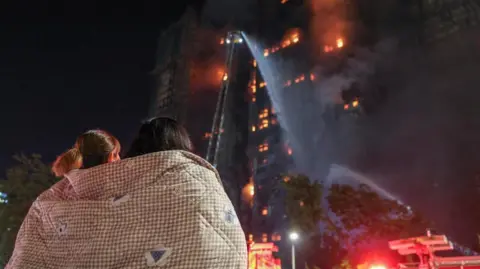 Reuters Young women wrapped in a blanket look up at burning high-rise blocks of flats in the darkness.  
