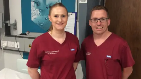NHS Humber Health Partnership A woman and a man standing side by side in a clinical room. They are wearing matching maroon NHS uniforms labelled "Advanced Clinical Practitioner". They are smiling, with hospital equipment and a bed in the background.