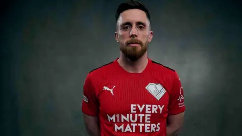 Sky Bet A man with short brown hair and a brown beard poses in red football kit against a black backdrop.