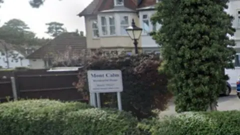 A leafy street with an old fashioned black lamppost, hedges , trees, a dark brown fence and a sign saying Mont Calm Residential Home in blue writing on a white background