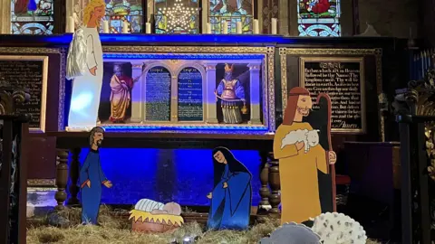 A large wooden cut-out nativity scene in front of a church altar - cartoon-style figures in traditional dress. It is colourful, with hay on the floor below a stained glass window.