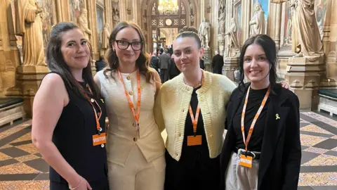 Four women stand in Westminster together smiling at the camera. They are dressed smart and have orange lanyards around their necks.