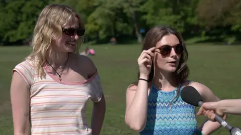 Two women in a park. On the left is a woman with blonde hair and sunglasses, wearing a white top with yellow and pink stripes. On the right is a woman with brown hair, wearing sunglasses and a blue top.