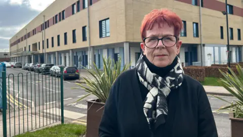 A woman with glasses, short read hair, a black and white scarf and dark top, stands outside a hospital building.