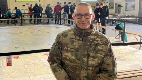 A man wearing a camouflage print jacket is standing indoors next to a large map of the moors. People are spread out around the room. 