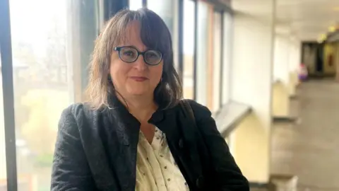 BBC Kirsty Chatwood looking straight at the camera. She is wearing a black jacket over a white shirt. She has dark hair and glasses. Her hands are folded in front of her. Her right arm is through the top of a silver crutch. She is standing in a stairwell in Cables Wynd House.