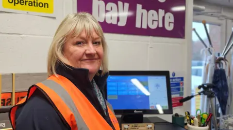 Paul Moseley/BBC Karen Betts is smiling at the camera, She's wearing an orange high-vis waist over a dark blue fleece. Behind her, we can see the till and a sign saying "pay here"