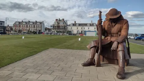 The Tommy statue shows a giant World War One soldier sat on a trunk. He is holding a gun. It is made out of brown metal and is next to the the green.