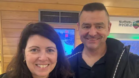 A couple smiling next to a Northern Ireland tourism banner. The female has black hair with gold hoop earrings. The male is wearing a black jacket his t-shirt button is opened, he is smiling showing his teeth.  