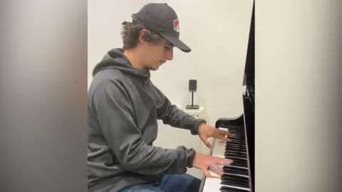 Brad Kella, usando um boné de beisebol, blusa de moletom e jeans tocando piano