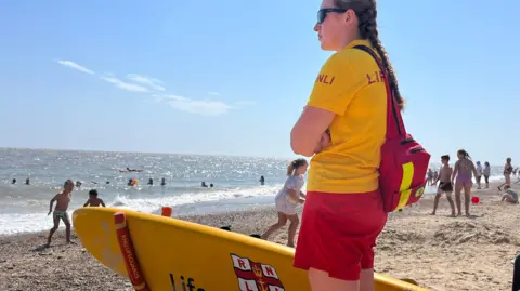 An RNLI lifeguard patrols Lowestoft south beach on a bright sunny day, 12 Aug 2024. She is wearing a yellow t-shirt,  red shorts and sunglasses and has a red bag over her right shoulder. She is standing beside an RNLI surfboard. Several children are running and playing in the background and there are people swimming in the sea in the distance.
