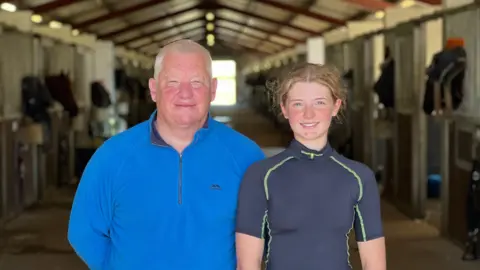 Kevin Frost (left) in a bright blue fleece and Lauren Frost (right) in a dark racing top facing the camera with stables stretching behind them