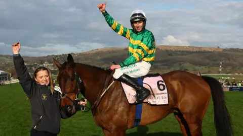 Reuters Mark Walsh riding a brown horse named Inothewayurthinkin. Mark is wearing a green and yellow striped top and looks at the camera with one hand raised in the air with his thumb up. A woman can be seen in the picture near the horse's head with her hand raised.