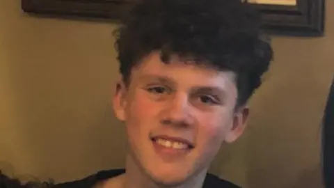 Family handout A teenage boy wearing a dark top. He has thick curly brown hair. He smiles at the camera. Behind him, there is a brown wall with the bottom of a picture frame visible.