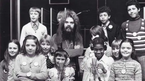 A group of young children wearing various badges gathered around a man with dark hair and a large beard