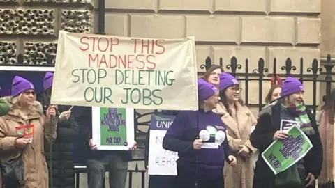 Protesters standing in a line wearing purple Unison hats. They are holding up signs and placards, the biggest of which reads: 'Stop the madness, stop deleting our jobs'. They are standing in front of black railings and a building.