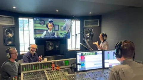 LizPatel Lauren Patel holding a figurine of PC Mukherjee, smiling in a studio. She is wearing a grey top and has long brown hair. 