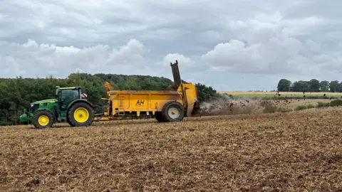 A green tractor is pulling a yellow trailer that is spreading muck on to a field of soil out the back of it.