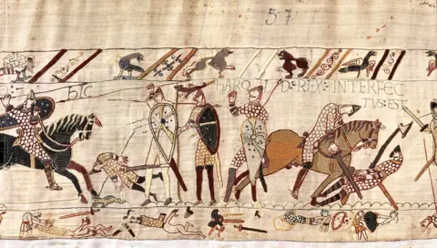 Bayeux Museum A wide crop shows the battlefield scene where Harold is depicted as being shot with an arrow surrounded by horses and knights, with birds and medieval writing reading 'Harold rex interfectvsest' with fabric detail showing the number 57.