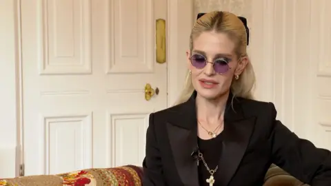 A woman with blond hair, tinted glasses and black outift sat on a sofa