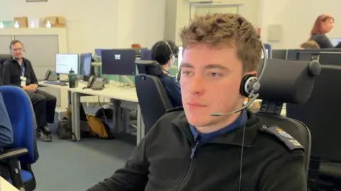 Hannah Roe / BBC A man wearing a black quarter zip with shoulder pads. He is sat in an office wearing a headset.