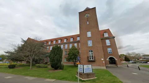 Google Spelthorne Borough Council. A brown brick building with a block tower. There is a patch of grass with trees in front of the building.