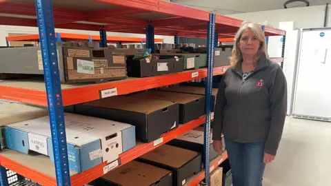 BBC A woman stands beside some shelves with upturned empty boxes on them