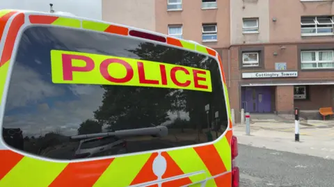 A West Yorkshire Police van with "POLICE" written in red on the back on a fluorescent yellow background. In the background of a shot is a red brick doorway with a sign saying COTTINGLEY TOWERS over it.