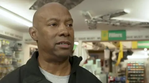 Neville Johnny inside his DIY shop. He wears a black jacket over a grey t-shirt.