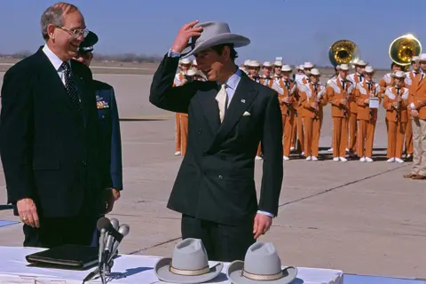 AP Charles dons a cowboy hat given to him by the mayor of Austin, Texas, during a visit to the city in 1986. A large brass band plays on the airport tarmac behind him