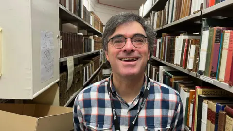 Ollie Conopo/BBC A smiling man in a check shirt and glasses stands in a corridor with lots of books on shelves on both sides of him.