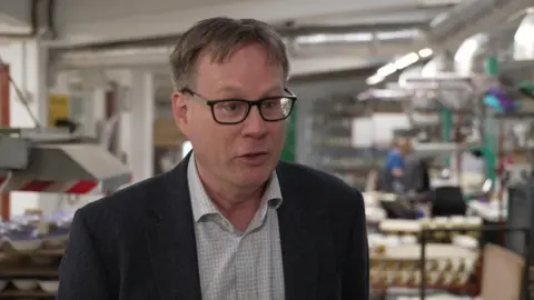A man wearing glasses, a shirt and a dark jacket is prominent in the photo. The background showing a workplace is blurred.