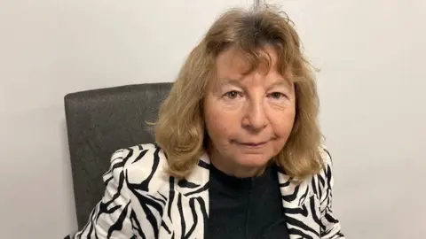 Eleanor Brazil, with shoulder-length hair, sitting in an office chair, wearing a black and white patterned jacket and dark top underneath